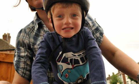 A close up of Karen and her son on a bike, he's sitting on a front-mounted bike seat and wearing a helmet, holding onto the handlebars. karen is leaning round to smile at the camera