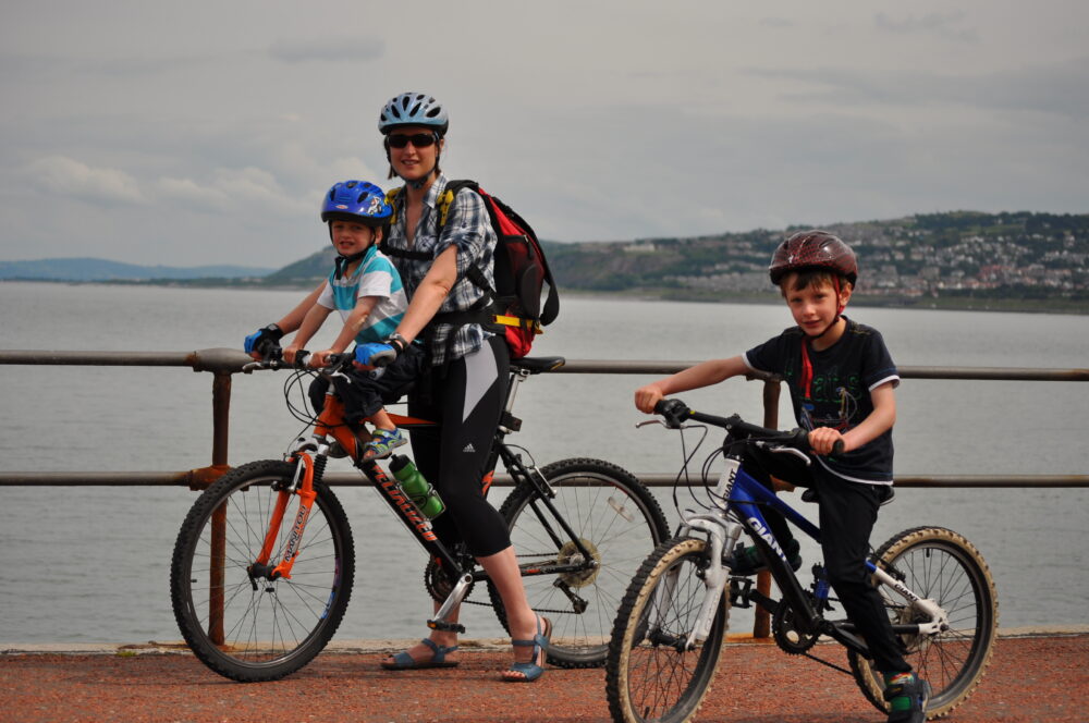 Karen Gee with her youngest son in a front bike seat, and her eldest on his own bike, by the coast