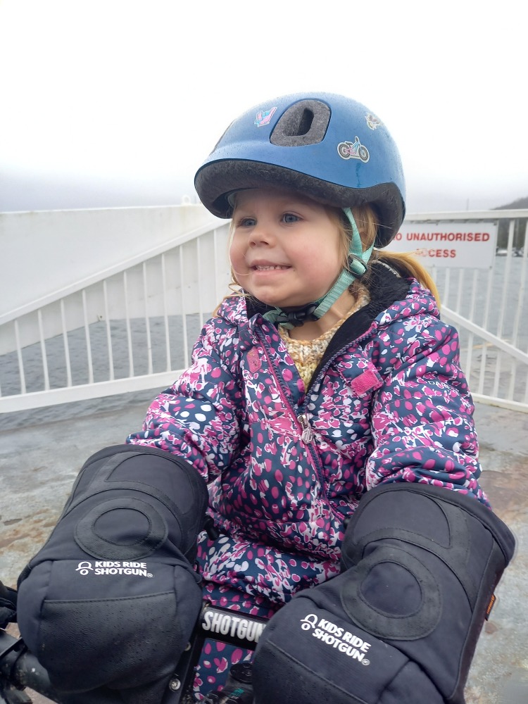 Girl in a blue helmet and flowery jacket using the Kids ride shotgun pogies while on a bike on board a boat.