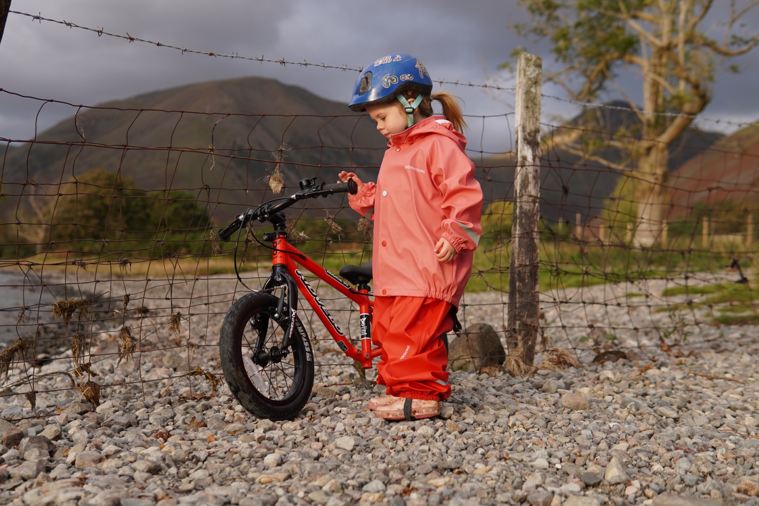 A girl in a pink coat stadning on a pebbly beach next to the kidvelo rookie 14 bike to review