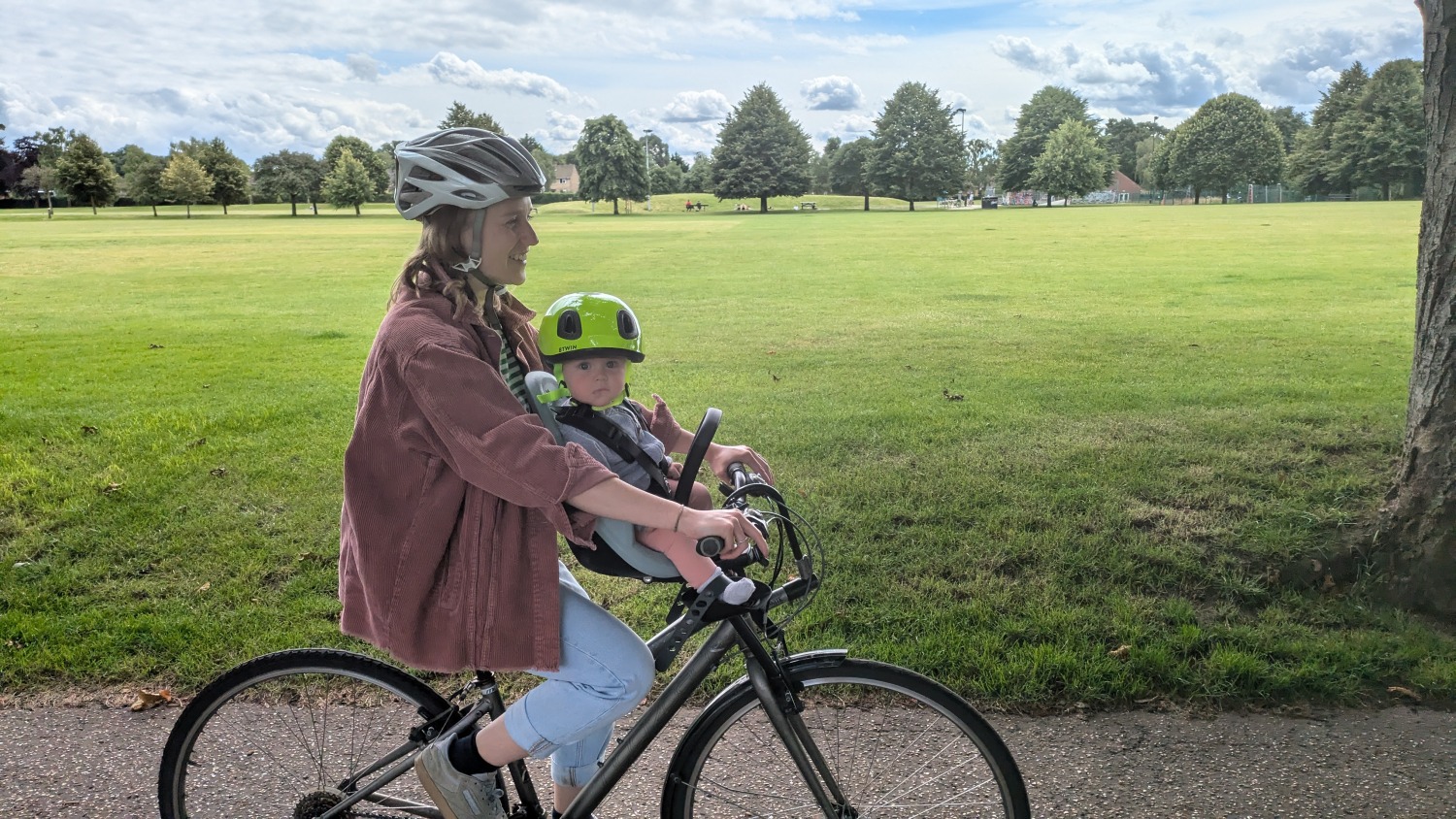 a mum in a pink shirt is riding in a park on a sunny day with her baby in a front thule yepp 2 mini seat