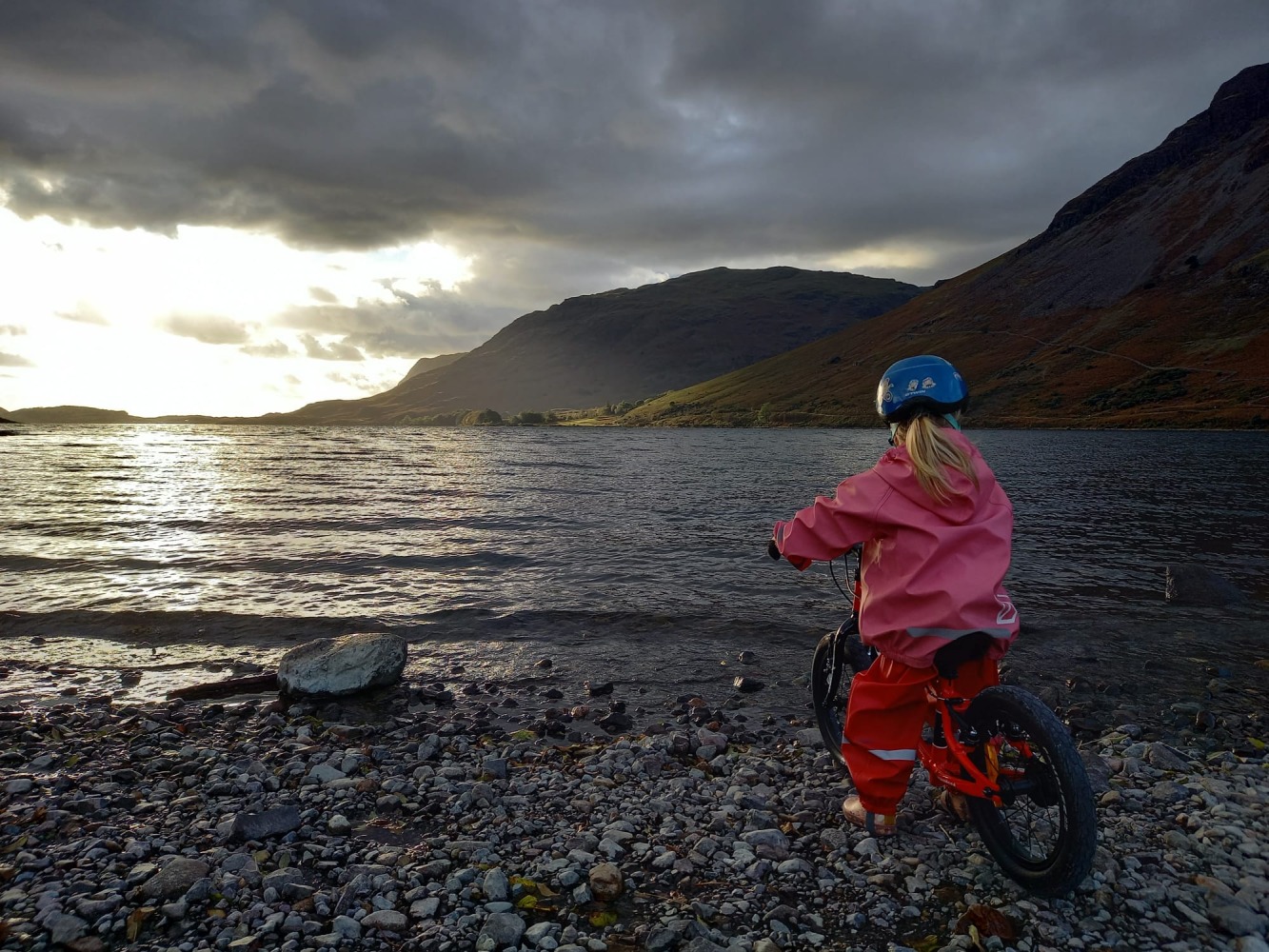 Best 14" kids' bikes: a little girl in waterproofs stands over her 14-inch bike next to the shoreline, looking out at sea with a mountain in background