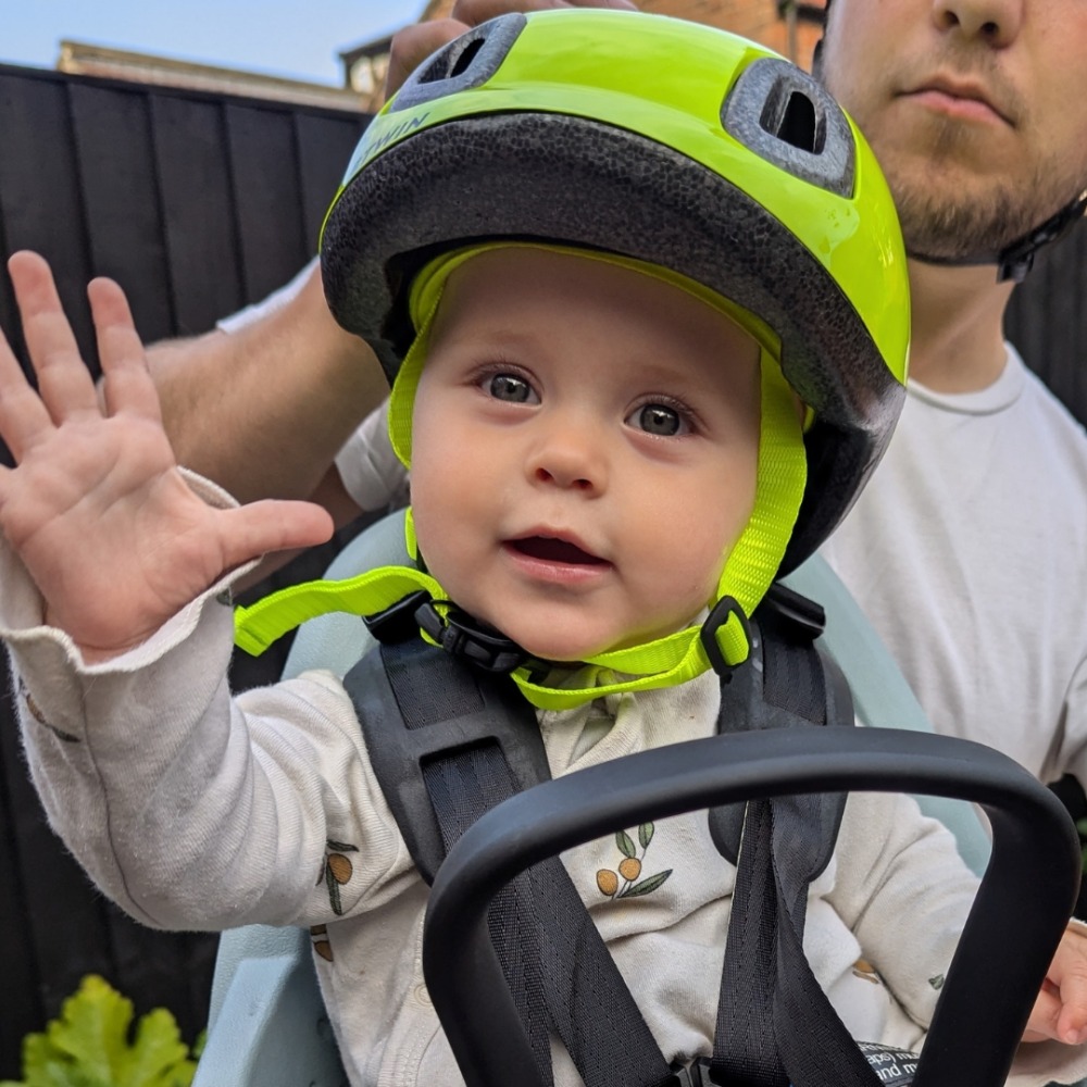 A baby girl wearing a brightly coloured bike helmet, sitting in a front-mounted bike seat and holding a hand up to the camera