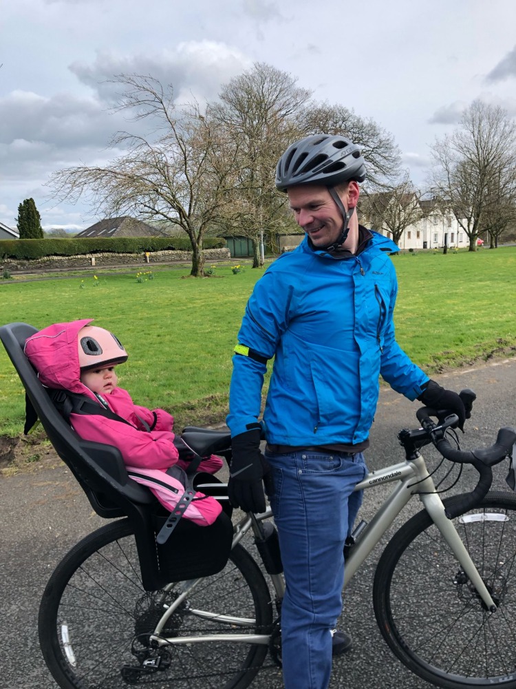 Thule Yepp 2 Maxi rear seat in use, a little girl in a pink onesie on the back of her parents bike with her dad smiling at her