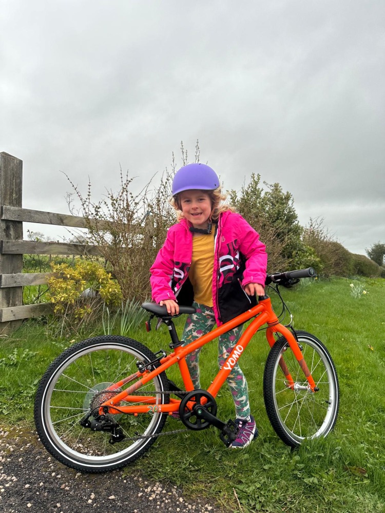 A girl in a pink jacket and purple helmet with her orange yomo 20 bike with a massive grin on her face