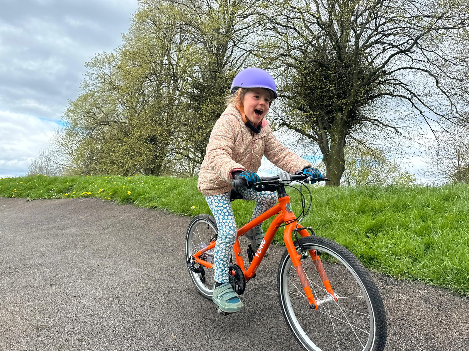 A 6 year old girl in a pink coat and purple helmet riding a orange yomo 20 bike on a pump track with a massive grin on her face