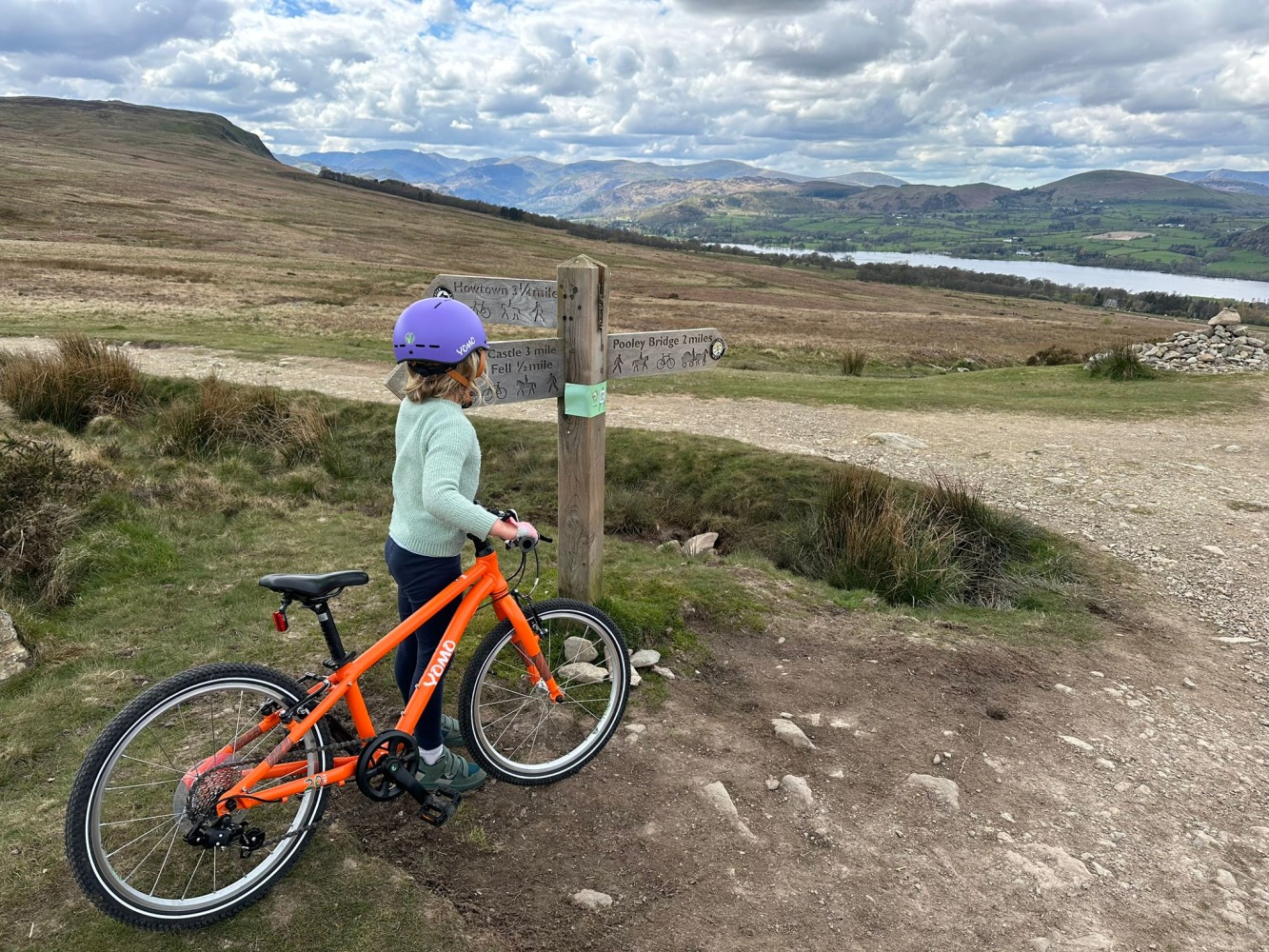 A girl in a green jumper is holding her orange yomo 20 bike pointing to a signpost that points over the fells overlooking a lake