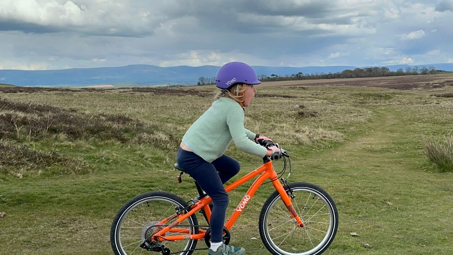 A 6 year old girl in a blue cardigan and purple helmet riding a orange yomo 20 bike on a grassy hill