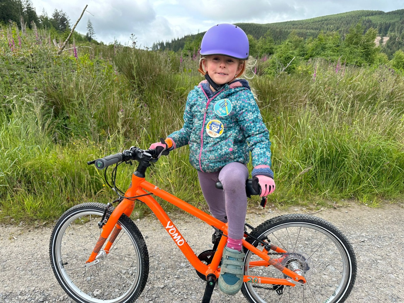 A 7 year old girl in a blue jacket and purple helmet riding a orange yomo 20 bike in the forest with a massive grin on her face