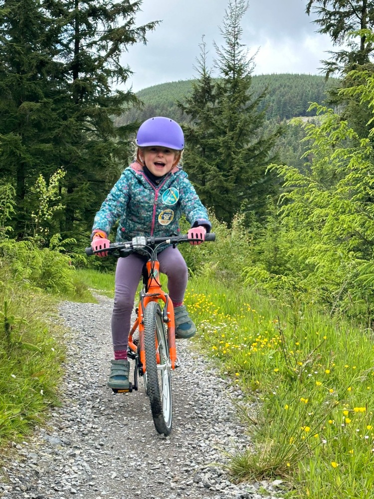 Best 20" kids' bikes: A 7 year old girl in a blue jacket and purple helmet riding a orange yomo 20 bike in the forest with a massive grin on her face
