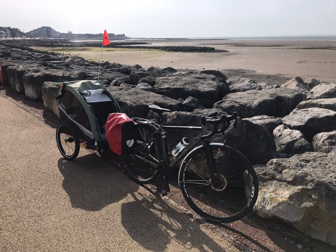 Burley D'Lite X child trailer hitched to an adult bike leaning against a rocky wall with a large sandy beach behind it.