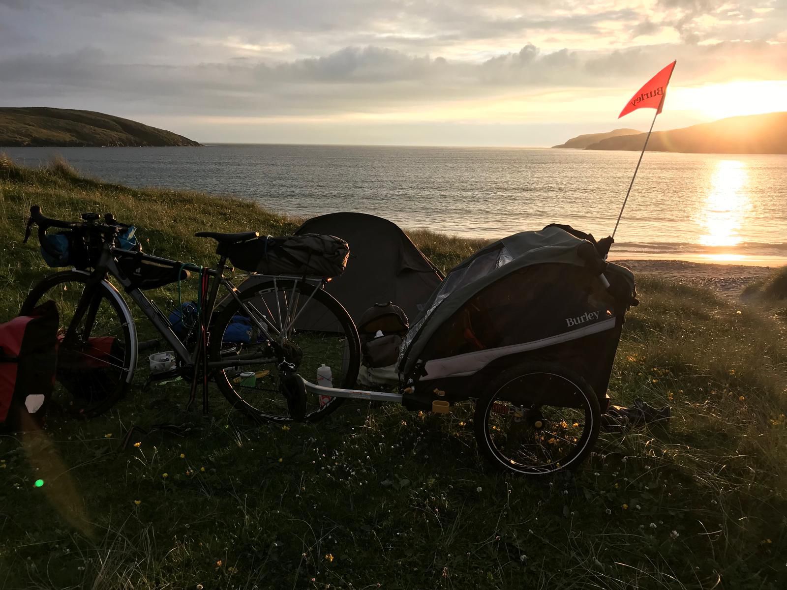 Burley D'Lite X child trailer hitched to an adult bike leaning against tent with the sun setting over the sea behind.