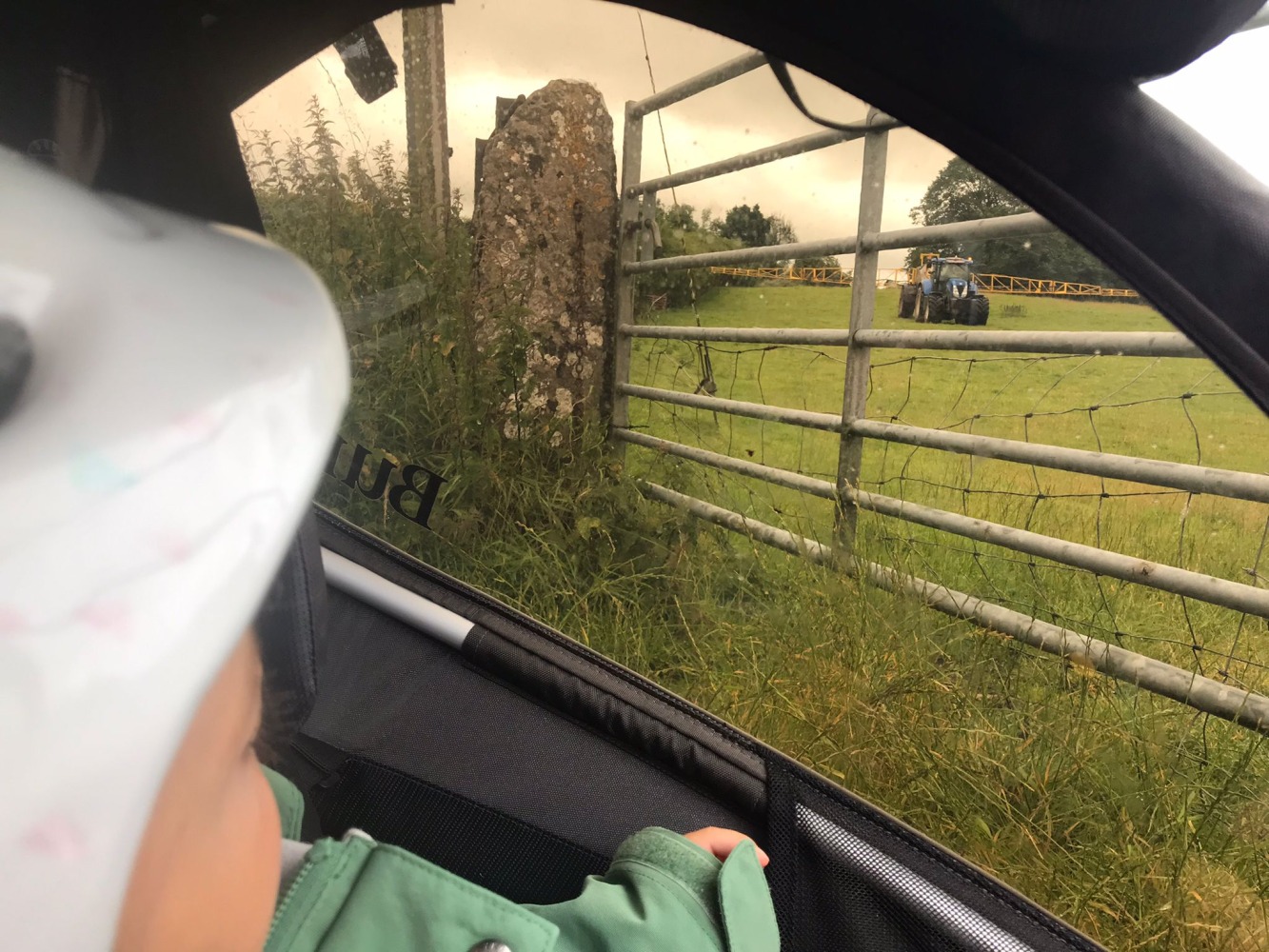 a little boy sitting in a burley D'Lite X trailer looking out of the window at a tractor on a field