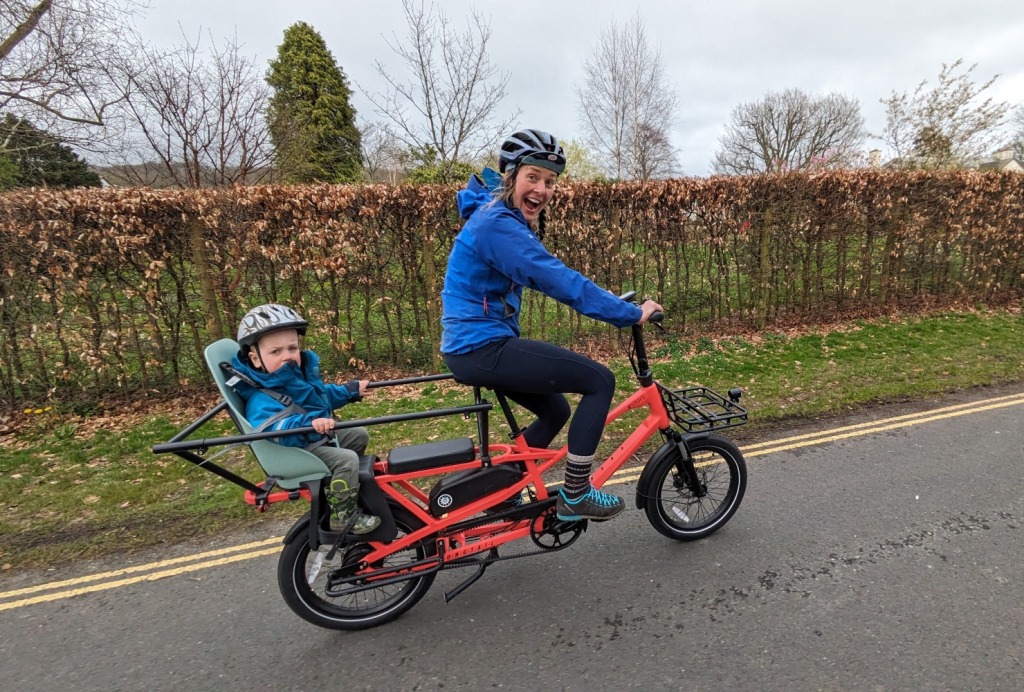 first family bike ride: A parent riding a long-tailed electric cargo bike with a child on the back, both smiling at the camera