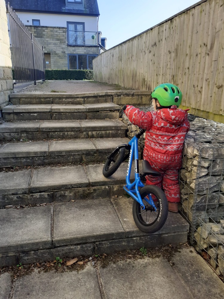 Kidvelo Rookie 12 review: a little girl in a red onesie carrying her blue balance bike up some stairs