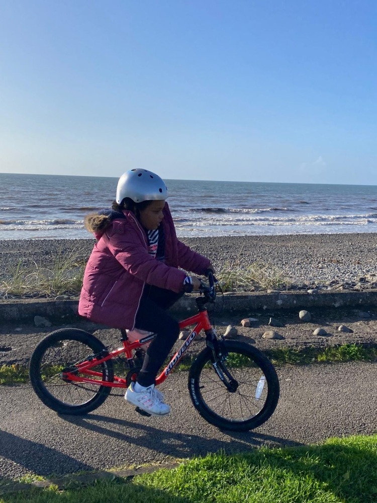 A black girl in a pink coat and silver helmet riding the kidvelo rookie 18 along the sea front