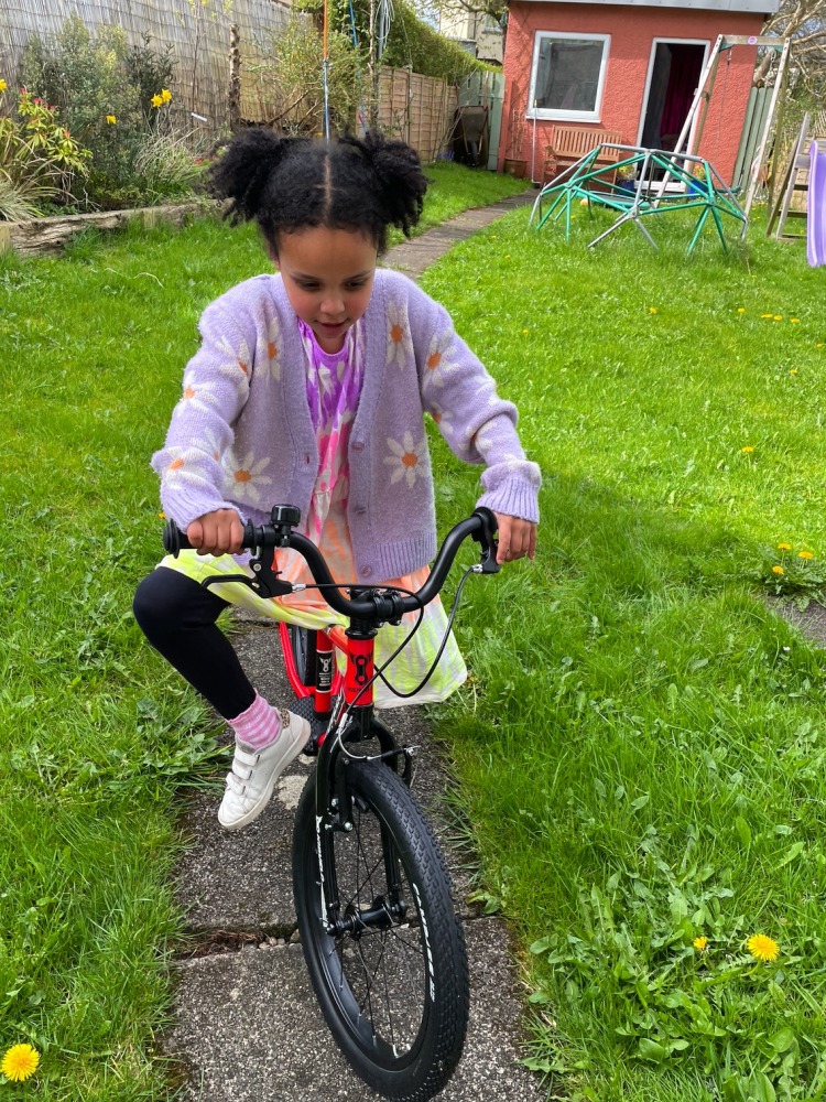 A black girl in a pink cardigan and bunches riding the kidvelo rookie 18 in her garden