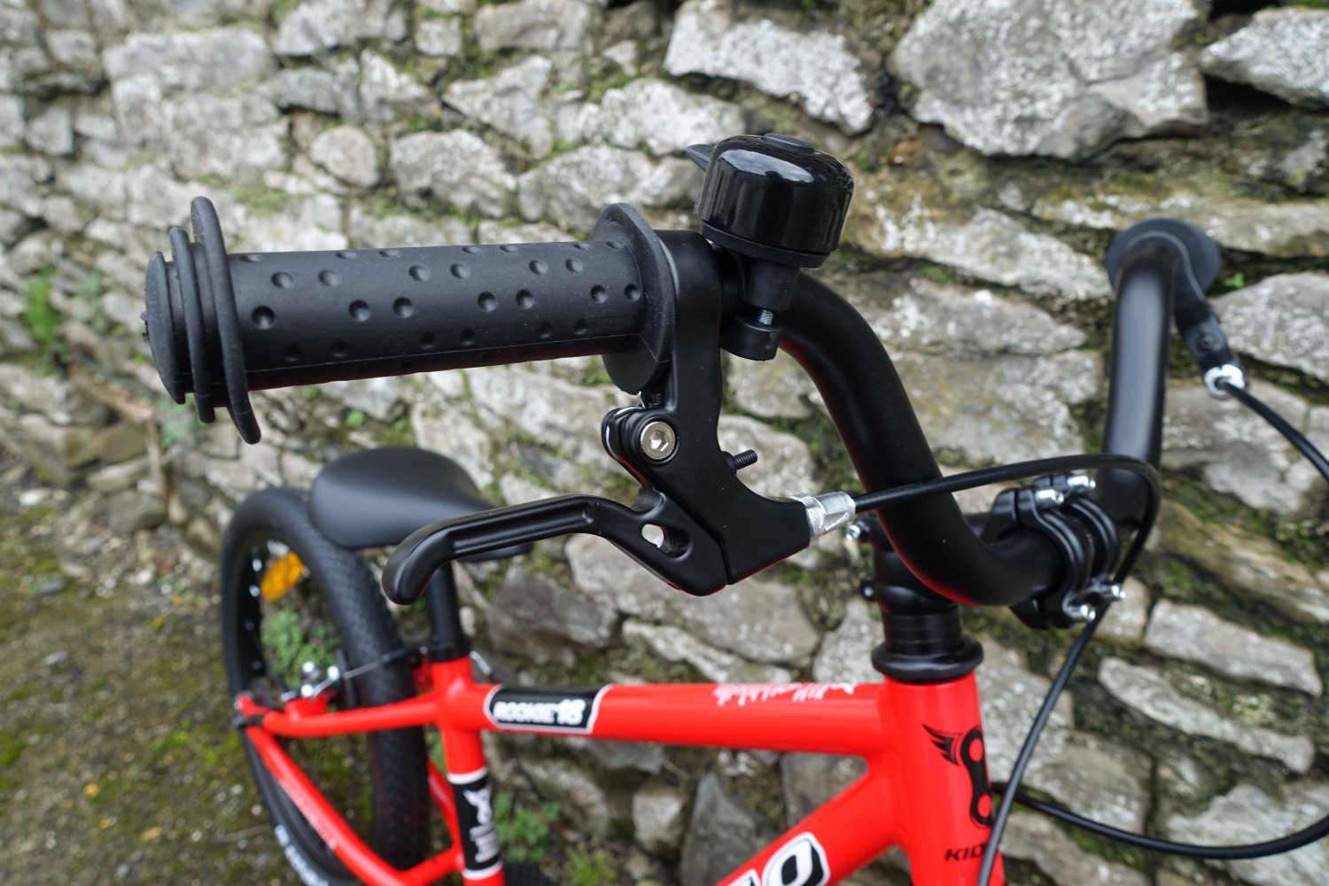 The kidvelo rookie 18 bike in red leant against a wall- a close up of the bell and brake lever