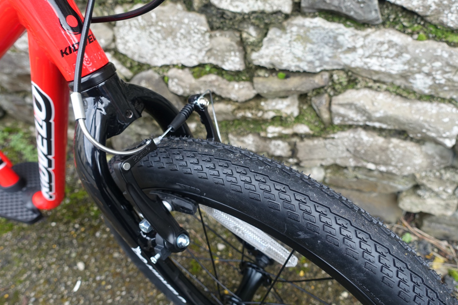 The kidvelo rookie 18 bike in red leant against a wall- a close up of tfront wheel and brake