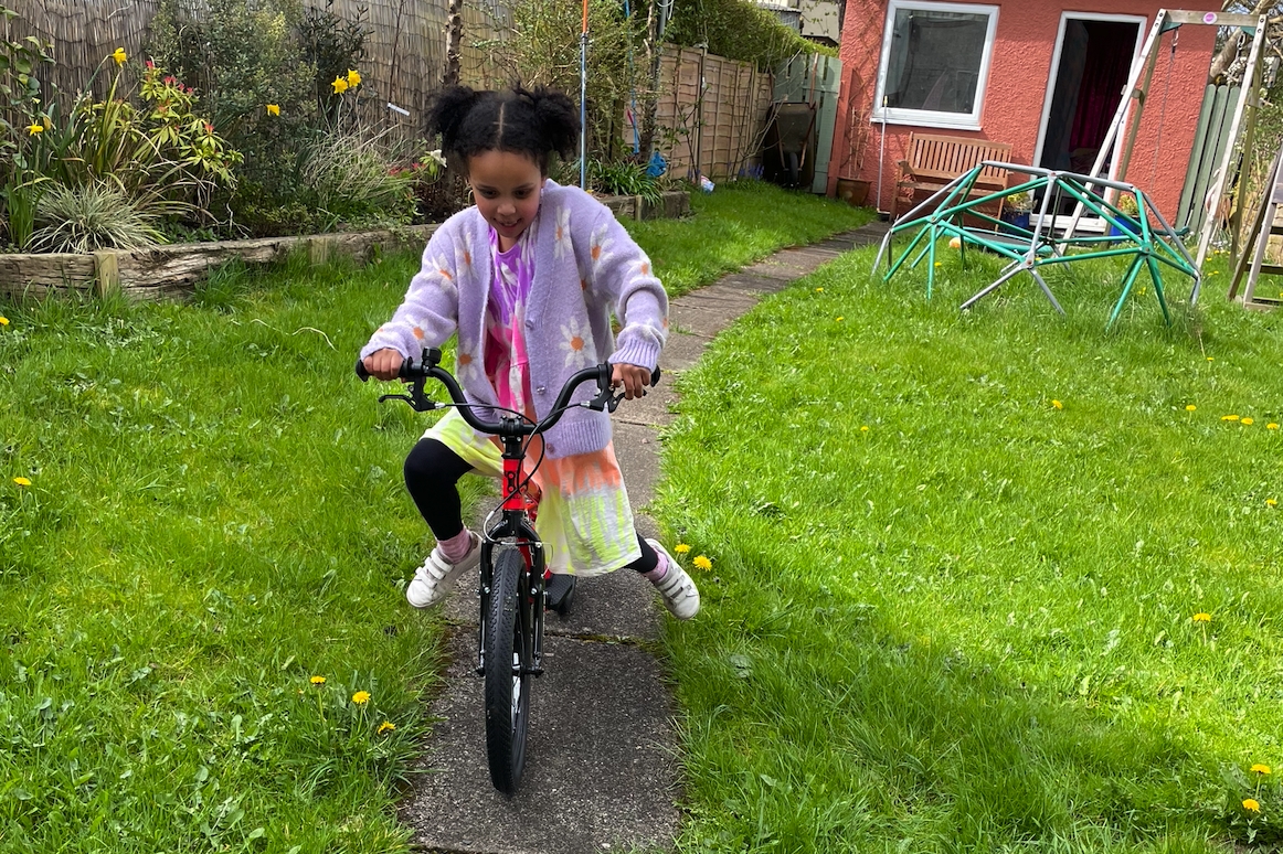 A girl in a pink cardigan and bunchies riding the kidvelo rookie 18 in her garden