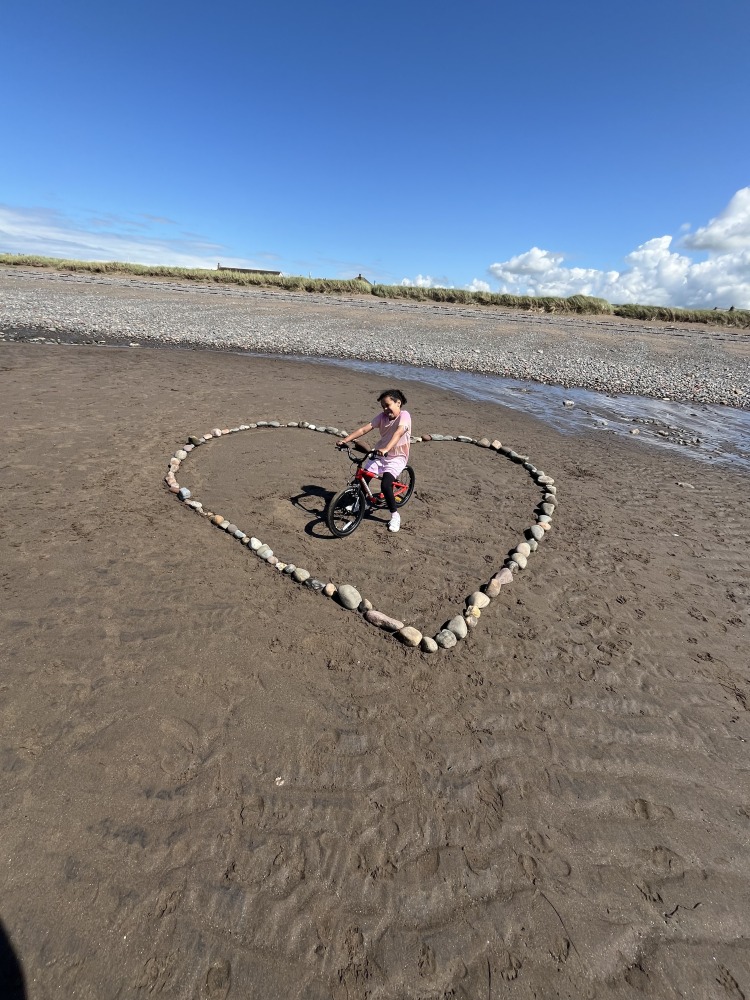 a black girl standing within a heart made of rocks on a beach on her kidvelo rookie 18 bike