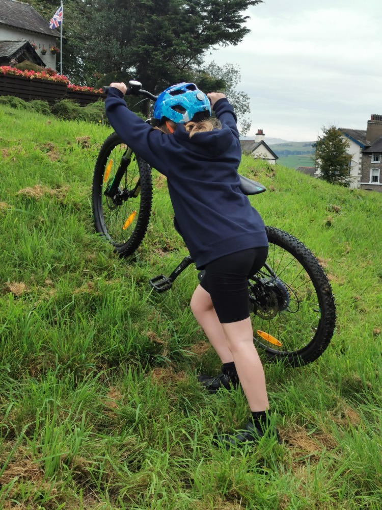 Cube acid 240 MTB review- a girl in a navy hoodie is pushing the cube acid mtb up a grassy slope