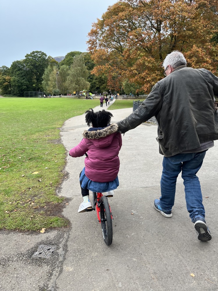 A black girl in a pink coat and afro hair riding the kidvelo rookie 18 in the park with her grandad holding on to her to keep her steady