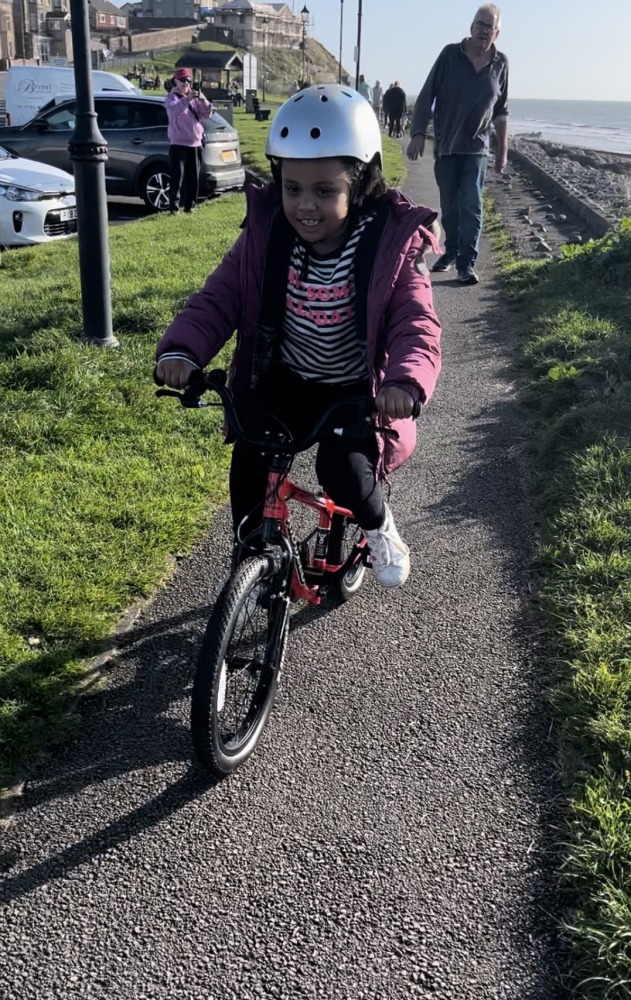 A black girl in a pink coat and silver helmet riding the kidvelo rookie 18 along the sea front