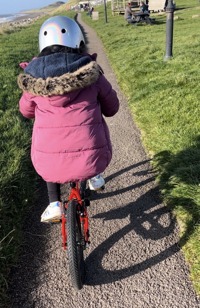 A black girl in a pink coat and silver helmet riding the kidvelo rookie 18 along the sea front