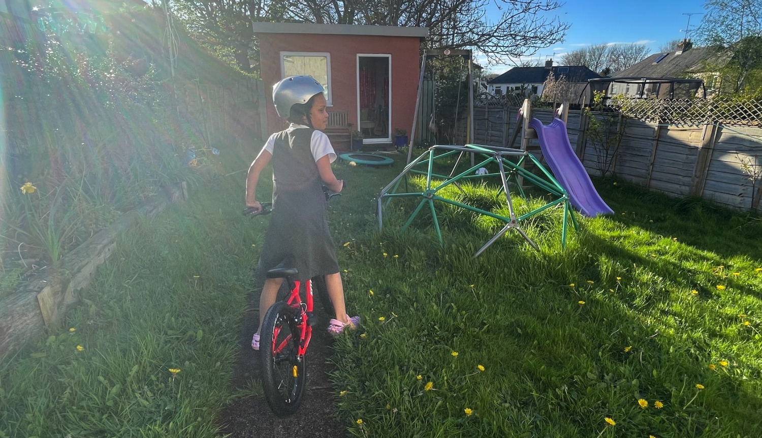 A black girl in her school dress and silver helmet riding the kidvelo rookie 18 in her garden