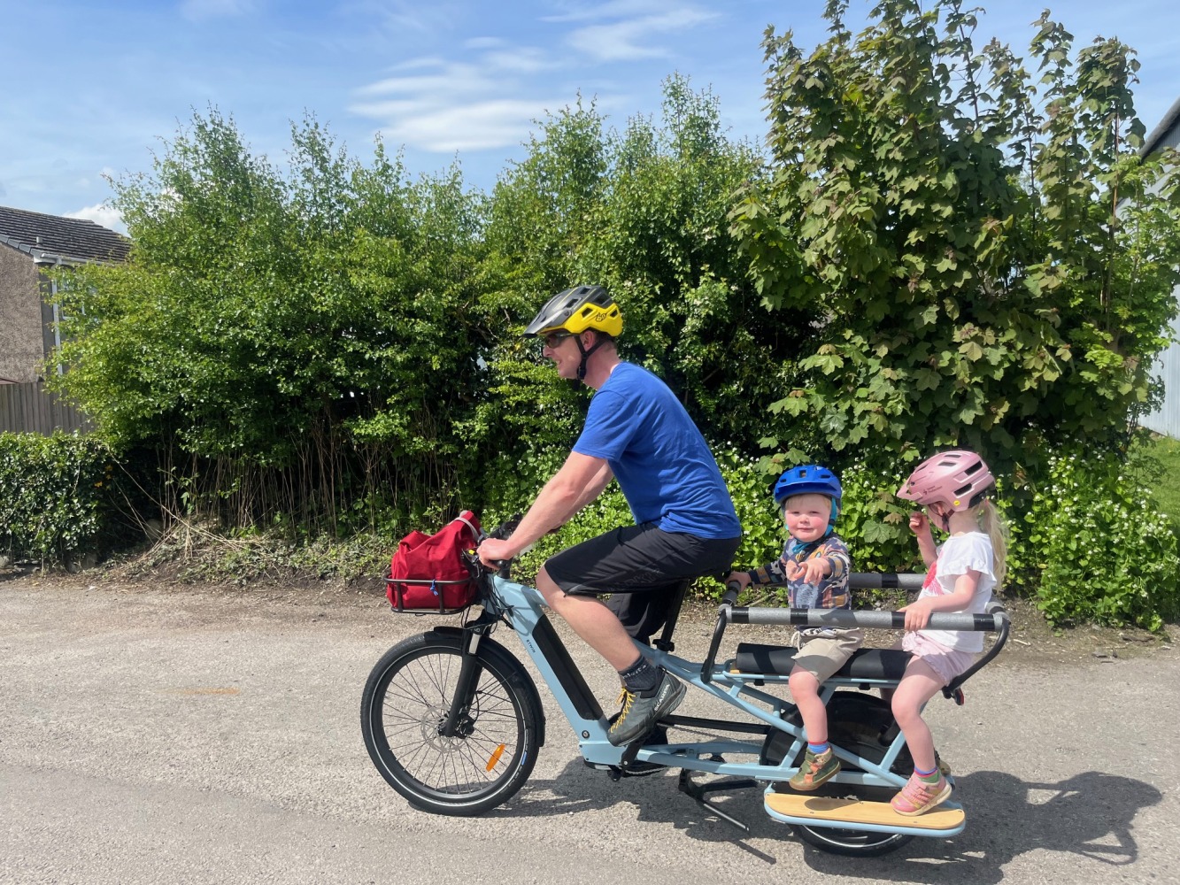 a family cycling on a summers day along a river, the two children are wearing colourful helmets and clothes and are looking at the camera while their dad cycles