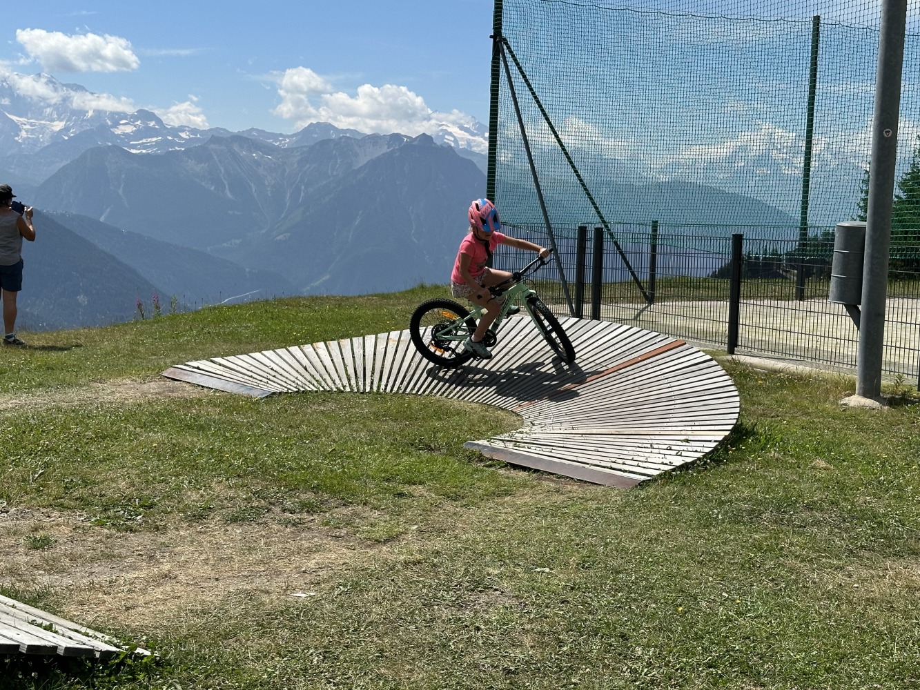 Specialized Riprock 20 review- a photo of a litle girl in a pink jacket riding the blue bike on a mountain bike trail in the alps- there is a beautiful mountain scene behind her