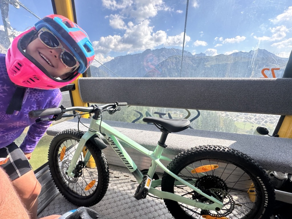 a very smily girl wearing sunglasses and a helmet riding a gondela in the alps with her blue specialized Riprock 20