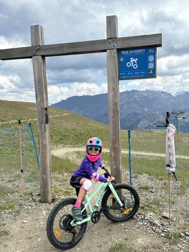 Specialized Riprock 20 review- a photo of a litle girl in a pink jacket riding the blue bike on a mountain bike trail in the alps- there is a beautiful mountain scene behind her