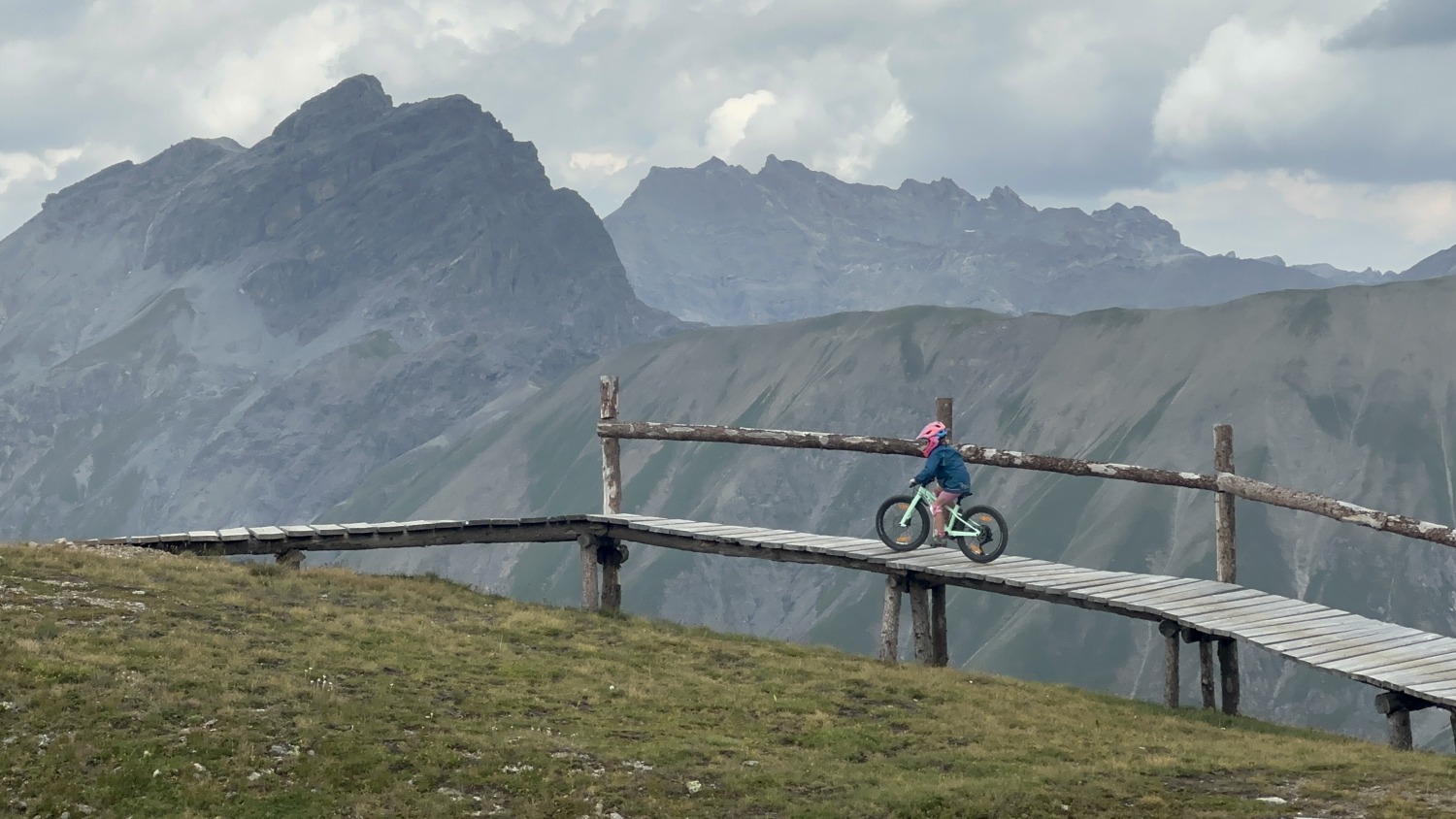 Specialized Riprock 20 review- a photo of a litle girl in a pink jacket riding the blue bike on a mountain bike trail in the alps- there is a beautiful mountain scene behind her