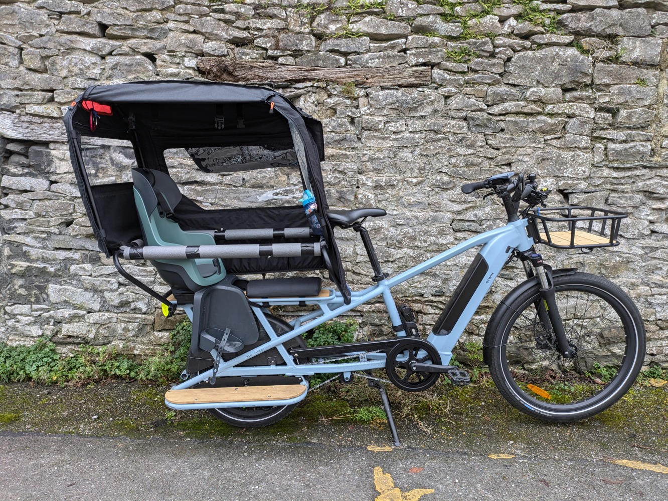 A light blue Decathlon longtail cargo bike with a rear rain cover on leant against a brick wall