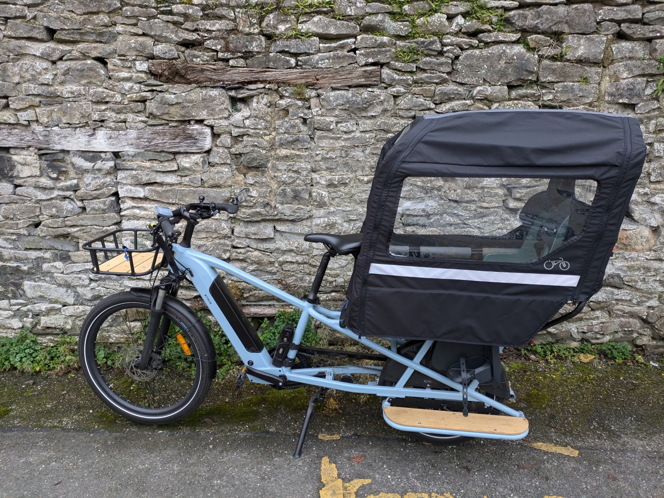 A light blue Decathlon longtail cargo bike with a rear rain cover on leant against a brick wall