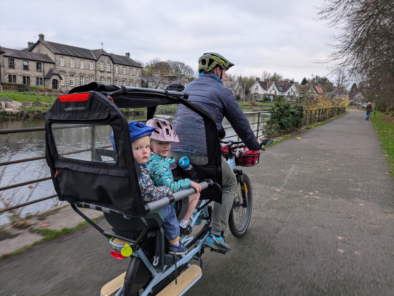 a family cycling by the river on a winter day along a river, the two children are wearing colourful helmets and clothes and are looking at the camera while their dad cycles