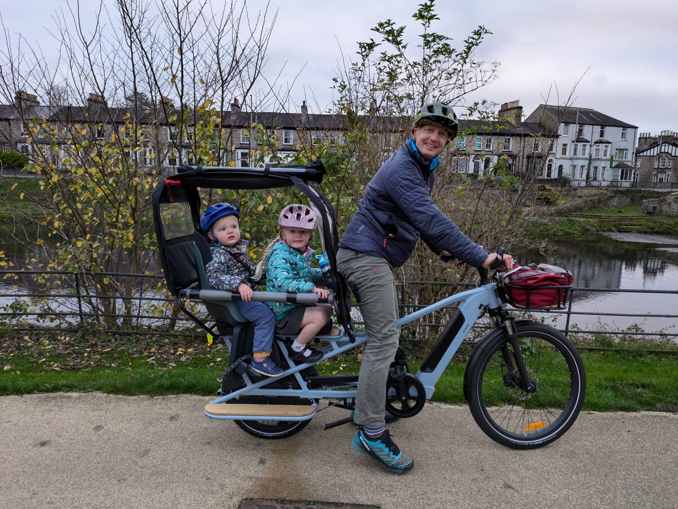 a family cycling by the river on a winter day along a river, the two children are wearing colourful helmets and clothes and are looking at the camera while their dad cycles