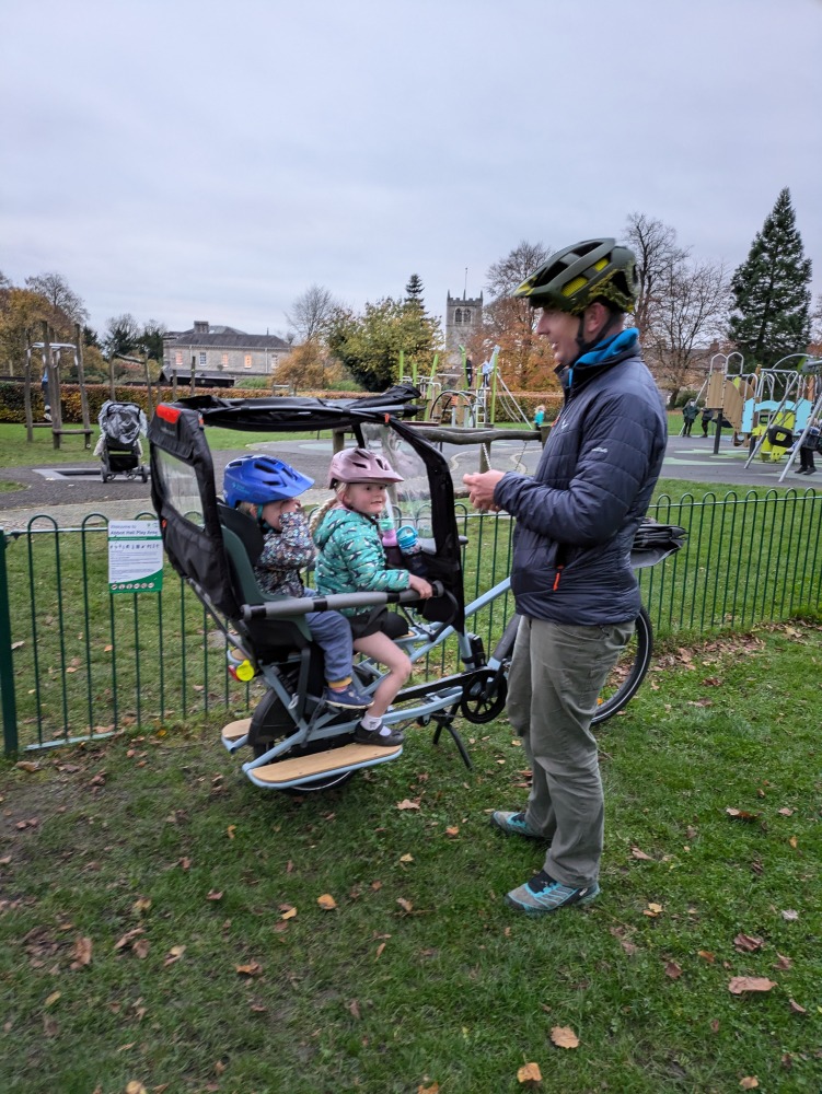 a dad giving his children snacks while they are sat on the back of a longtail cargo bike