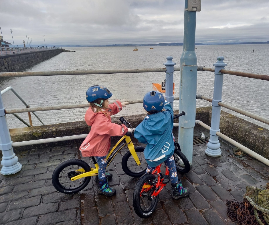 Two girls on 14" wheel balance bikes pointing at a sign next to the sea