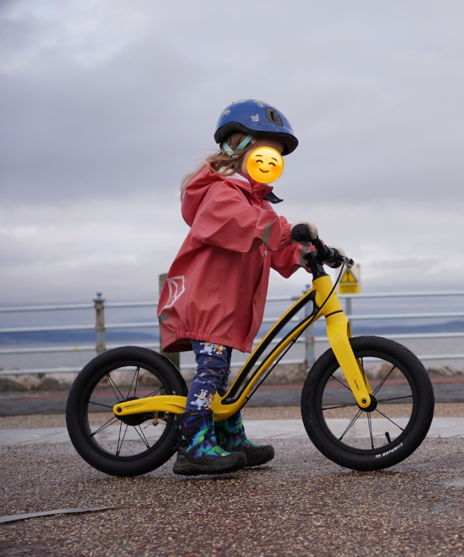 Yellow Hornit AIRO 14 balance bike being ridden by a girl in a pink jacket through a puddle