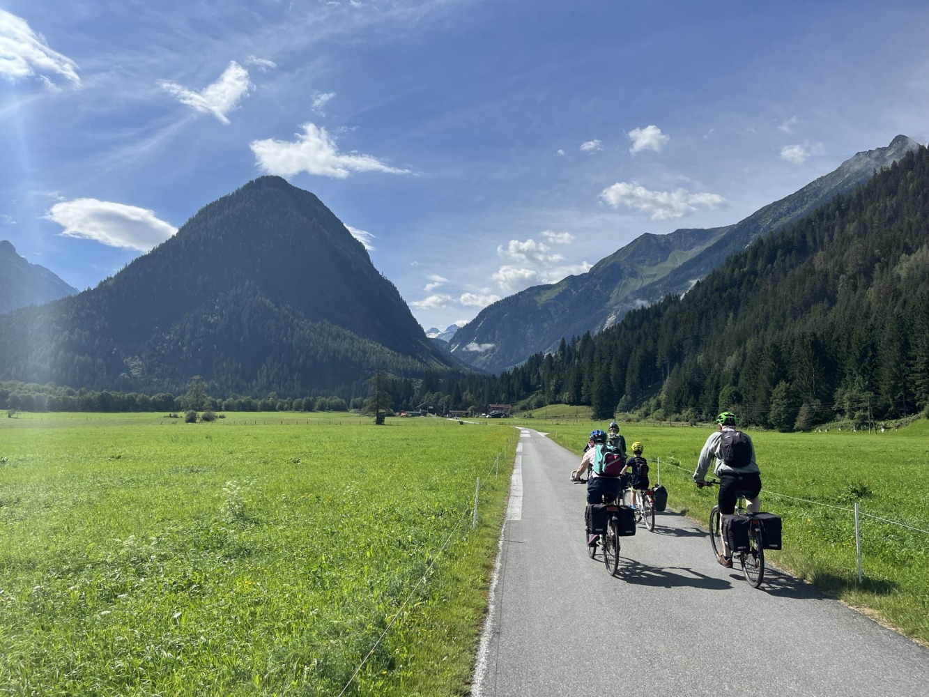 Bea's partner, children and mother seen cycling from behind along a smooth paved path through a green field and mountains in the distance