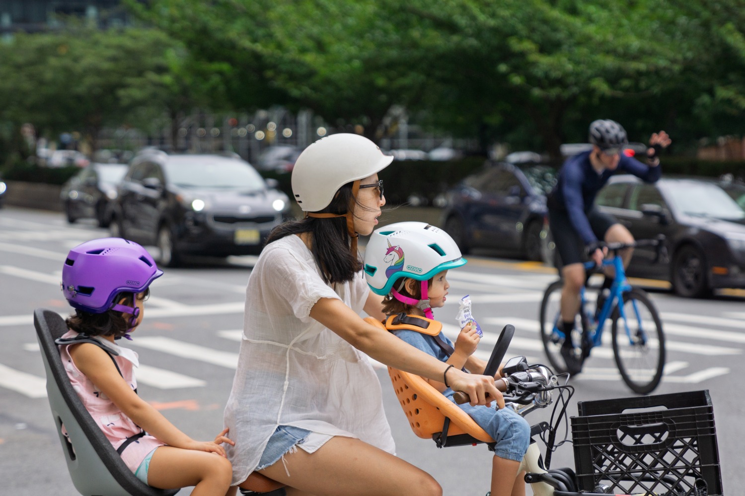 An Asian woman cycling a bike with her toddler on a front-mounted bike seat, wearing an Ozmo helmet, and a small child on a rear-mounted bike seat, also wearing an Ozmo helmet.